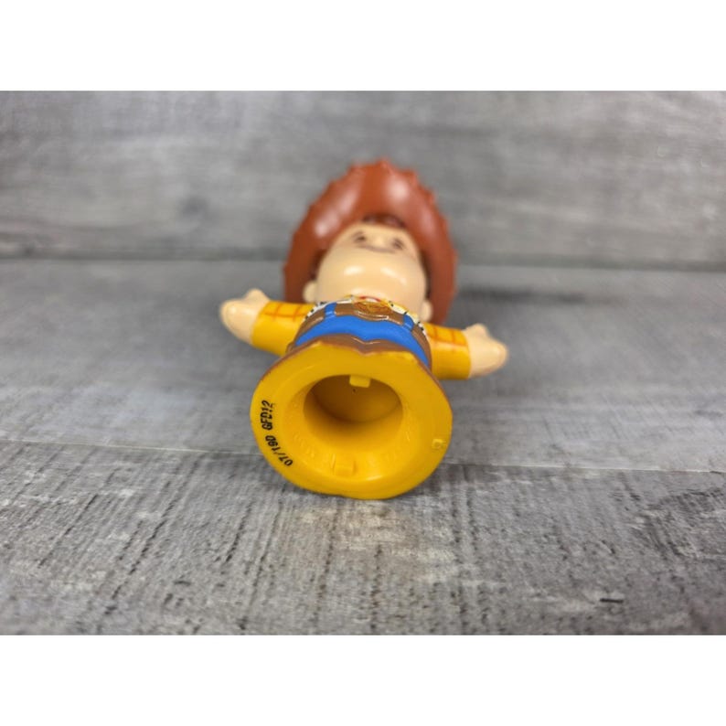 May include: A yellow, blue, and brown toy figure of a cowboy character with outstretched arms. The figure has a brown cowboy hat, a yellow shirt, and blue pants. The figure's base is yellow with the text "07/12" and "GFD12".