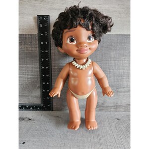 Pu&ograve; includere: Una bambola di plastica marrone con capelli neri ricci, che indossa una collana di conchiglie bianche e un pannolino di stoffa marrone. La bambola &egrave; in piedi su una superficie di legno.