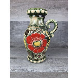May include: A ceramic pitcher featuring a prominent embossed red flower with a yellow center, surrounded by green and brown foliage. The vessel has a cream-colored base, a decorative beaded rim, and a curved handle with a green and cream glaze.