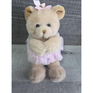 Pu&ograve; includere: Un orsacchiotto di peluche con una pelliccia color marrone chiaro, che indossa una gonna di tulle rosa e un fiocco rosa sulla testa. L'orso ha occhi neri e un piccolo naso marrone. Le braccia dell'orso sono incrociate davanti al petto.