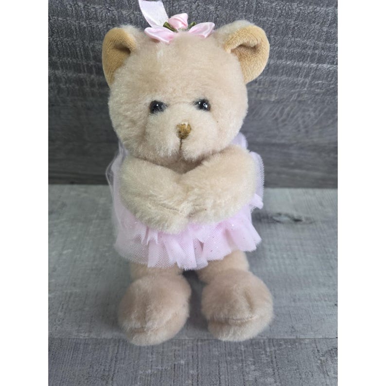 Pu&ograve; includere: Un orsacchiotto di peluche con una pelliccia color marrone chiaro, che indossa una gonna di tulle rosa e un fiocco rosa sulla testa. L'orso ha occhi neri e un piccolo naso marrone. Le braccia dell'orso sono incrociate davanti al petto.