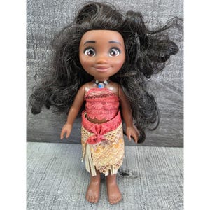 Op de afbeelding: Pop van een Polynesisch meisje met een donkere huid, bruine ogen en lang zwart haar. Ze draagt een rood en oranje top, een gele en oranje rok en een blauw-witte ketting. De pop staat op een grijs oppervlak.
