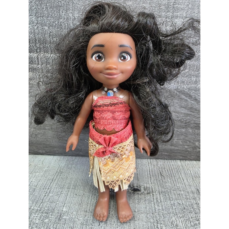Op de afbeelding: Pop van een Polynesisch meisje met een donkere huid, bruine ogen en lang zwart haar. Ze draagt een rood en oranje top, een gele en oranje rok en een blauw-witte ketting. De pop staat op een grijs oppervlak.