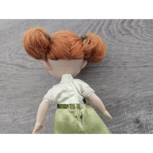 Puede incluir: Una mu&ntilde;eca de pelo rojo que lleva una camisa blanca y una falda verde brillante. La mu&ntilde;eca tiene una etiqueta en la parte posterior de la cabeza que dice "Hecho en China".