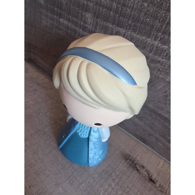 May include: A figurine of a character with light blonde hair, a blue headband, and a blue dress. The figurine has a round head and a stylized design. The figure is standing on a surface with a wood grain pattern.