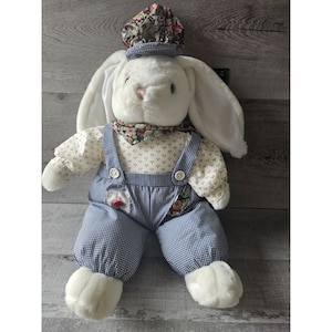 May include: A white plush bunny wearing blue and white checkered overalls, a floral print shirt, and a floral print hat. The bunny has long white ears and a pink nose.