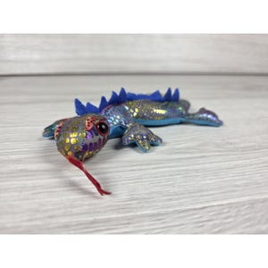 May include: A plush lizard toy featuring a metallic purple and gold scale pattern, large dark eyes, and a long red felt tongue. The creature has a row of blue fabric spikes running along its back and four splayed legs with detailed feet.