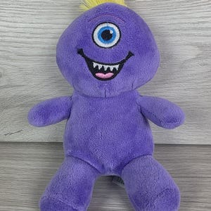 May include: A purple plush monster toy featuring a single large blue eye, a wide black mouth with visible white teeth, and a small tuft of yellow hair on top of its round head. The soft fabric body has two arms and two legs in a seated position.