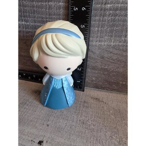 May include: A small, stylized figurine of a character with blonde hair and a blue dress. The figurine has a light blue headband and a blue dress with decorative patterns. The figure is approximately 4 inches tall.