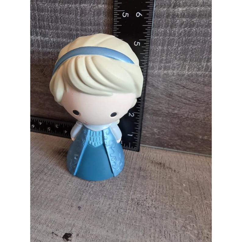 May include: A small, stylized figurine of a character with blonde hair and a blue dress. The figurine has a light blue headband and a blue dress with decorative patterns. The figure is approximately 4 inches tall.
