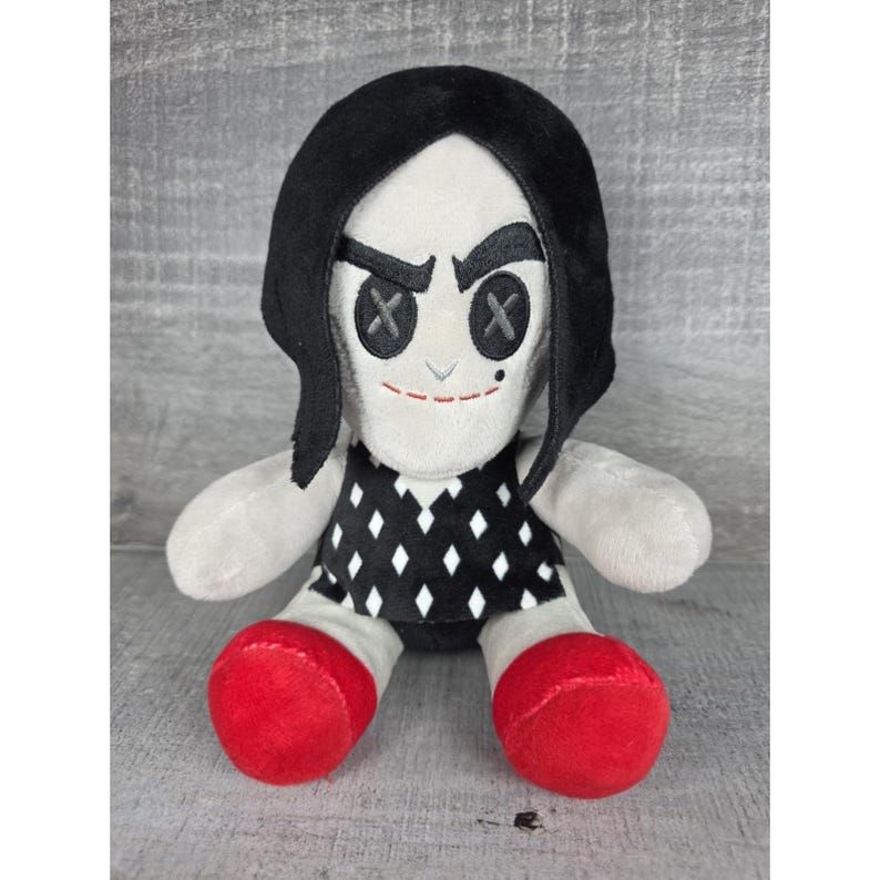 May include: A plush doll with a white face, black hair, and a black and white diamond pattern dress. The doll has button eyes, a stitched smile, and red shoes. The doll is a soft toy.