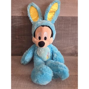 May include: A plush Mickey Mouse toy dressed in a light blue bunny costume. The costume features large, upright ears with yellow polka dots. Mickey's face is visible, with his signature black nose and smiling expression. The toy is soft and cuddly.