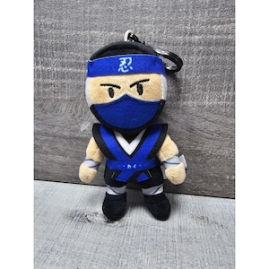 May include: Plush ninja keychain featuring a blue headband with Japanese characters, a blue mask, and a blue and black outfit. The plush toy has tan arms and a black keychain attached to the top.
