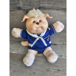 Puede incluir: Un perro de peluche que lleva un traje de marinero azul con un cuello blanco y una cinta roja, blanca y azul. El perro tiene una cara blanca y marrón con ojos marrones y un mechón de pelo blanco en la cabeza.