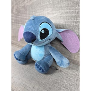 May include: A plush toy character with blue fur, a light blue chest patch, and large, pointed ears with light purple inner sections. The character features a dark blue nose, embroidered black and white eyes, and a small tuft of fur on its head.