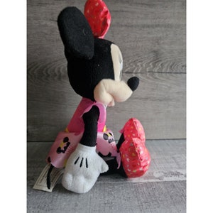 May include: A plush Minnie Mouse toy wearing a pink dress with white polka dots and red shoes with white polka dots. The toy is sitting on a wooden surface.