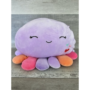 Puede incluir: Un pulpo de peluche morado con una cara sonriente y corazones coloridos en la cabeza. El pulpo tiene tentáculos de colores arcoíris.