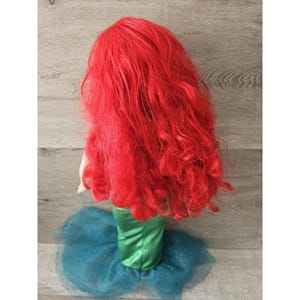 Puede incluir: Una mu&ntilde;eca sirena de pelo rojo con cola verde y falda de tul azul. La mu&ntilde;eca tiene el pelo largo y rizado y lleva un disfraz verde y azul.