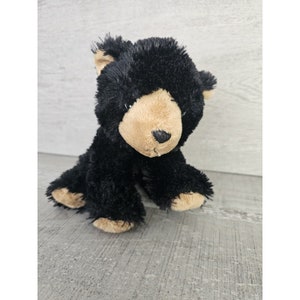 May include: A black plush bear with a brown face and nose. The bear is sitting on a light gray surface.