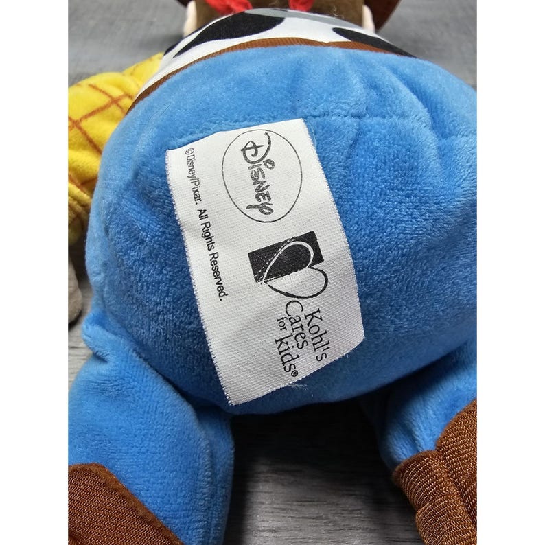 May include: A plush toy of the character Woody from the movie Toy Story. The toy is wearing a cowboy hat, a yellow shirt with a brown pattern, and blue pants. The Disney and Kohl's Cares for Kids labels are visible.