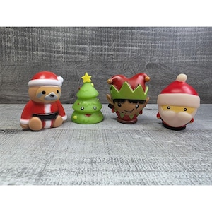 May include: Four festive Christmas figurines: a sloth in a Santa outfit, a green Christmas tree with a yellow star, an elf, and Santa Claus. The figures are brightly coloured and sit on a grey wooden surface.