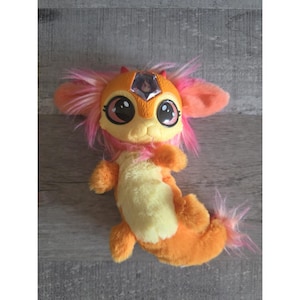 Puede incluir: Un peluche con forma de criatura de colores vibrantes. Presenta tonos naranja, amarillo y rosa, con grandes ojos expresivos y una gema en la frente. El peluche tiene orejas prominentes y una cola tupida, con un diseño fantasioso.
