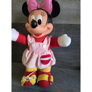 May include: A Minnie Mouse plush doll with a pink bow, black ears, and a red and pink outfit. The doll has white gloves, red shoes with yellow accents, and a pink dress with a red heart-shaped pocket.
