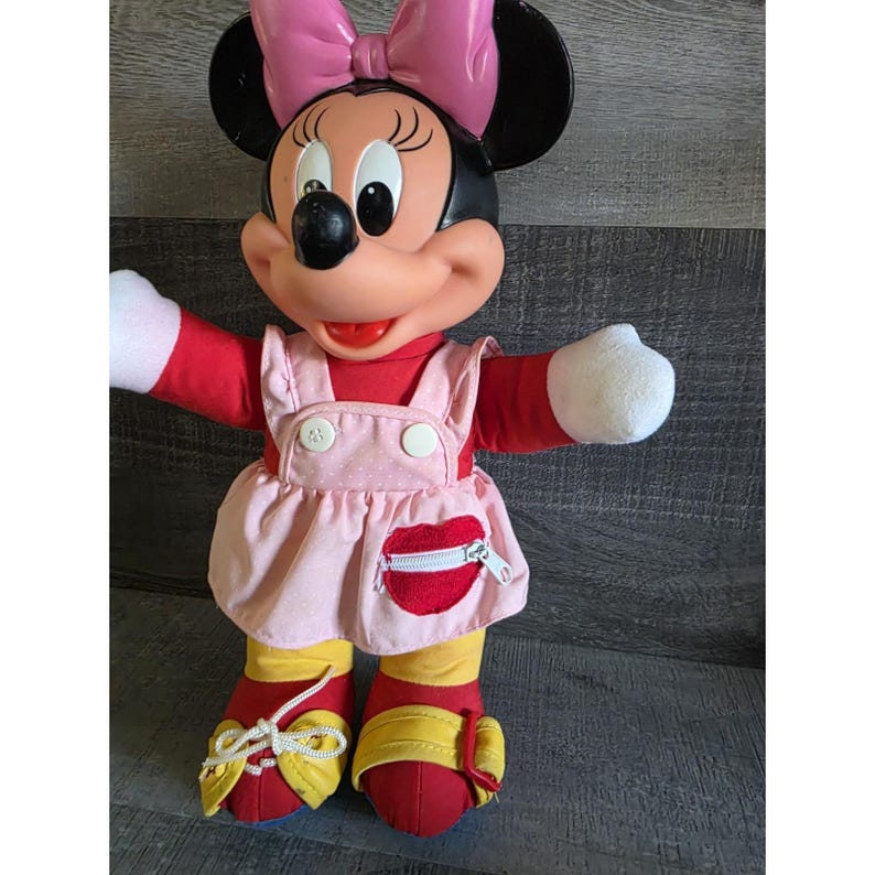 May include: A Minnie Mouse plush doll with a pink bow, black ears, and a red and pink outfit. The doll has white gloves, red shoes with yellow accents, and a pink dress with a red heart-shaped pocket.