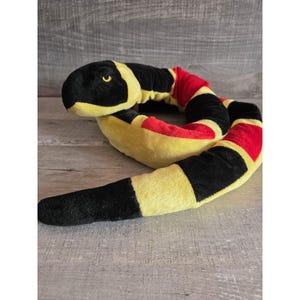 May include: A plush snake toy in black, yellow, and red. The snake's head is black with yellow eyes. The body is segmented with alternating black, yellow, and red sections. The toy is soft and appears to be made of fabric.