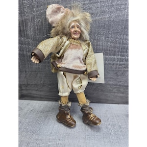 Könnte beinhalten: Eine skurrile Puppenfigur, bekleidet mit einer gold-braunen Jacke, cremefarbenen Shorts und braunen Stiefeln. Die Puppe hat ein lächelndes Gesicht, graues Haar und einen passenden Hut. Die Puppe ist ein dekorativer Artikel.