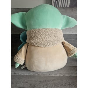 May include: A plush toy of a green character with large ears and a brown fur coat. The toy is sitting on a brown surface.