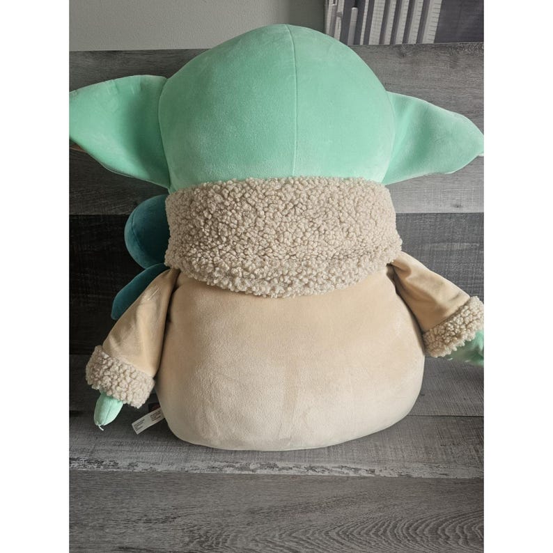 May include: A plush toy of a green character with large ears and a brown fur coat. The toy is sitting on a brown surface.