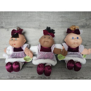 Può includere: Tre bambole Cabbage Patch Kids che indossano abiti bianchi con orlo in pelliccia e corpetti in velluto bordeaux con pois bianchi. Le bambole hanno diverse tonalità di pelle e indossano fiocchi bordeaux abbinati nei capelli. Ogni bambola ha un'etichetta con il suo nome e la data di nascita.
