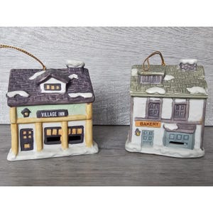 May include: Two ceramic Christmas village houses with snow-covered roofs. The house on the left is a "Village Inn" with a green awning and yellow trim. The house on the right is a "Bakery" with a brown awning and white trim.