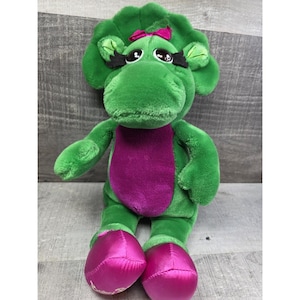 Könnte beinhalten: Ein grünes Dinosaurier-Plüschtier mit einem lila Bauch und rosa Füßen. Das Spielzeug hat eine rosa Schleife auf dem Kopf und schwarze Wimpern. Das Spielzeug befindet sich auf einer Holzoberfläche.