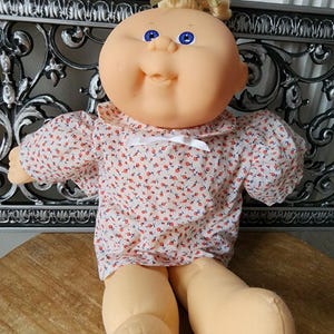 May include: A doll with a round, peach-toned face, bright blue eyes, and blonde hair tied with a gold ribbon. The doll wears a white shirt featuring a small red floral pattern, a rounded collar, and a thin white ribbon bow at the neckline.