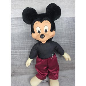 May include: A vintage Mickey Mouse plush doll. The doll has a black head and ears, a tan face, and a black shirt. It wears maroon satin pants and has white gloves and shoes. The doll is a classic Disney collectible.