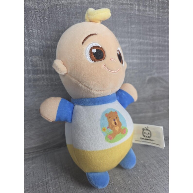 May include: A soft toy doll depicting a baby-like character with fair skin, wearing blue and yellow clothing, and a small tuft of yellow hair. The doll has a smiling face and a bear graphic on the front.
