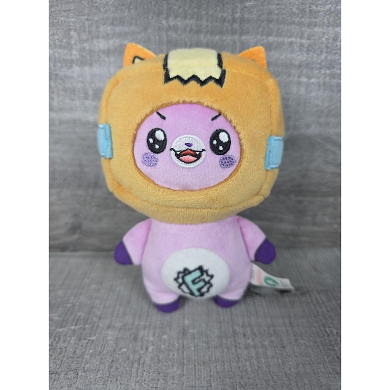 May include: A plush toy featuring a light purple character wearing a tan, box-shaped headpiece with orange ears. The character has embroidered black eyes, a small mouth with visible teeth, and a white circular patch on its chest with a blue letter F.