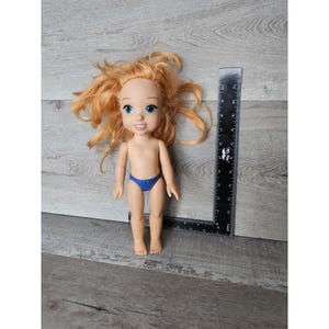 May include: A red-haired doll with blue eyes and a blue swimsuit. The doll is approximately 30 cm tall and is standing on a wooden surface.