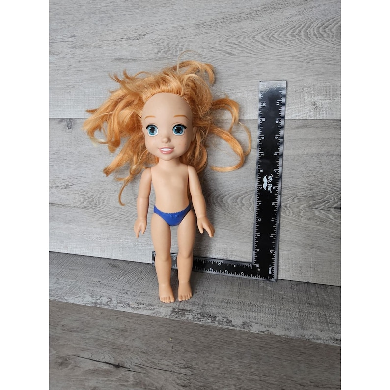 May include: A red-haired doll with blue eyes and a blue swimsuit. The doll is approximately 30 cm tall and is standing on a wooden surface.