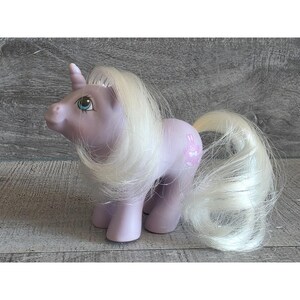 Puede incluir: Un juguete de unicornio de color lavanda con melena y cola blancas. El unicornio tiene ojos azules y un gráfico de conejito rosa en un costado. El cuerno es blanco con detalles en rosa. Un juguete coleccionable vintage.