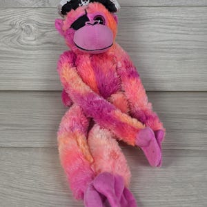 May include: A plush monkey toy featuring vibrant pink and orange tie-dye fur, a purple face, and long limbs. The monkey wears a black pirate hat with a white skull and crossbones emblem and a black eye patch over one eye, with soft purple feet.