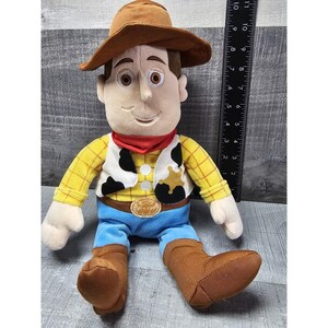 May include: A plush toy of a cartoon cowboy character, featuring a brown hat, a red neckerchief, a yellow plaid shirt, a black and white vest, blue pants, and brown boots. The toy is approximately 10 inches tall.