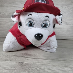 May include: A plush toy of a white Dalmatian dog character with black spots, wearing a red vest and a matching red baseball cap. The character has large embroidered blue eyes, a black nose, and floppy ears. The soft fabric construction allows the toy to lay flat in a pillow-like shape, featuring the Paw Patrol logo on the front of the cap.