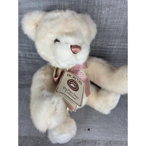 May include: A cream-colored teddy bear from The Boyds Collection, with a pink ribbon around its neck and a tag. The bear has a brown nose and a friendly expression. The tag reads "The Patty Duke Collection."