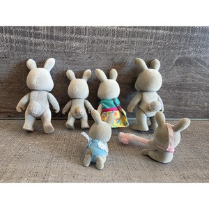 Puede incluir: Seis figuras de conejos grises en varias poses. Un conejo lleva un atuendo azul, otro un vestido colorido y uno tiene un accesorio rosa. Los conejos tienen colas peque&ntilde;as y redondas y orejas largas, sobre un fondo de vetas de madera gris.