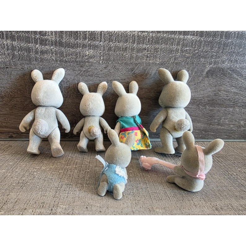 Puede incluir: Seis figuras de conejos grises en varias poses. Un conejo lleva un atuendo azul, otro un vestido colorido y uno tiene un accesorio rosa. Los conejos tienen colas peque&ntilde;as y redondas y orejas largas, sobre un fondo de vetas de madera gris.