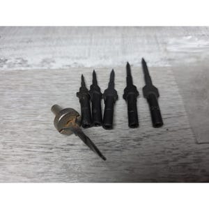 May include: A collection of black metal tools with pointed tips, arranged on a light-colored wooden surface. One tool has a silver-colored head. The tools appear to be for a specific purpose, possibly related to crafting or repair.