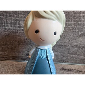 May include: A figurine of a cartoon character with blonde hair and a blue dress. The figure has a smiling face and is standing upright. The dress has a detailed design.